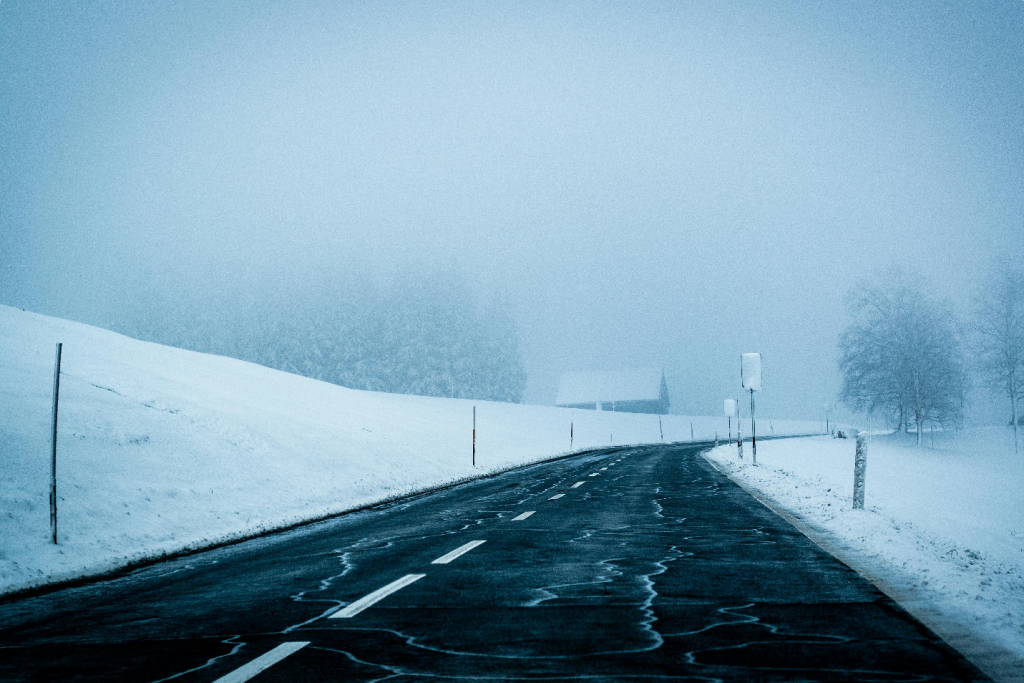 A road captured during snowfall, a winter hazard for St. Louis drivers, showing ice on the surface and snow on either side with the faint silhouette of bare trees visible in the distance